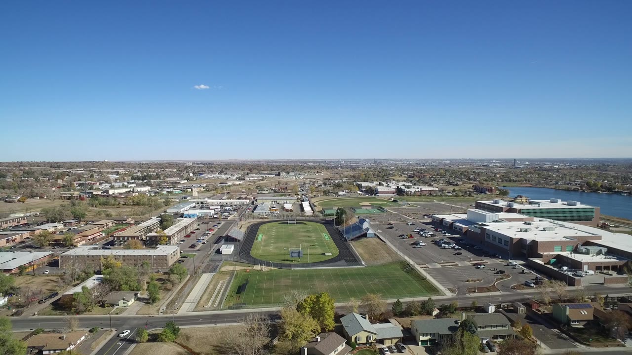 Wide drone view of a football field, commercial buildings, neighborhood streets, and water in the distance.