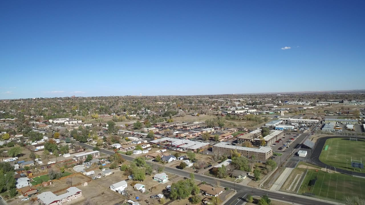 Wide aerial frame over Hidden Lake, Colorado showing residential blocks, larger buildings, parking lots, a green athletic field, and a flat horizon with no visible people.