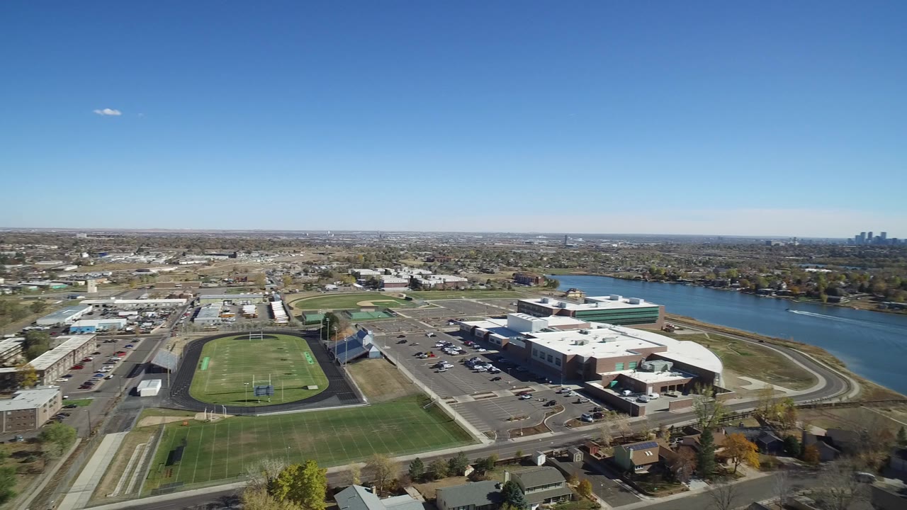 Aerial frame over Hidden Lake, Colorado showing ball fields, a track, low buildings, parking lots, houses, a broad waterway, distant skyline, and no visible people.