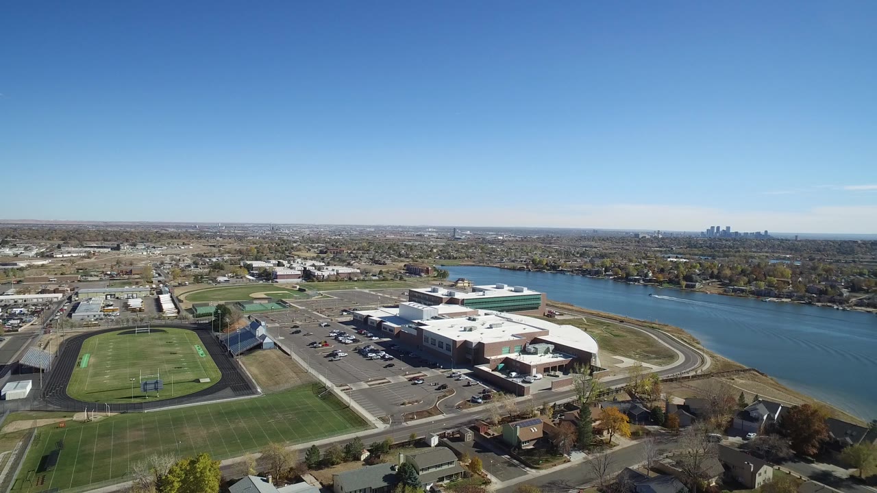 Drone-height view of fields, large buildings, parking lots, houses, and river-like water.