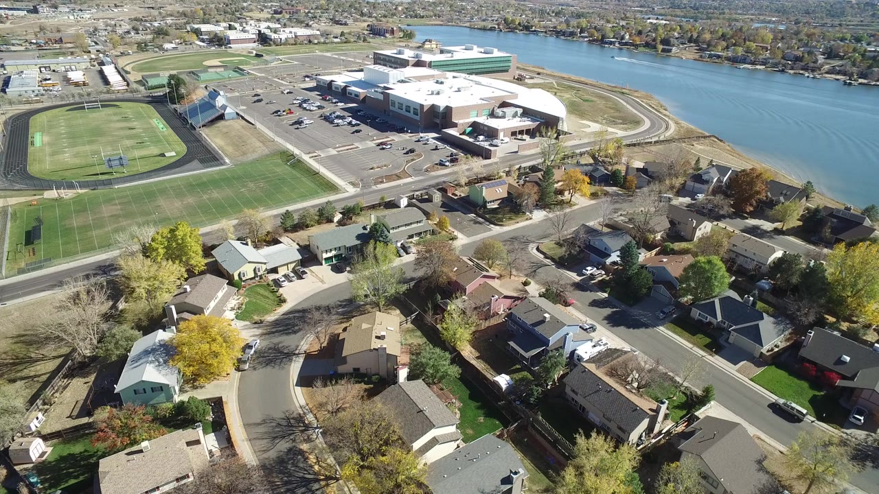 Opening aerial frame over Hidden Lake, Colorado homes with a green sports field and running track at left, a large building near the water, autumn trees, and no visible people.