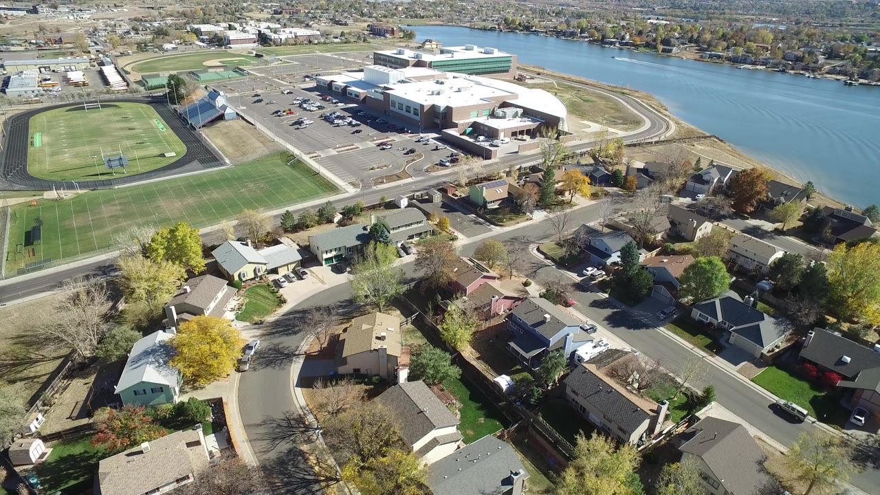 Second aerial frame in Hidden Lake, Colorado showing foreground rooftops, a curved residential street, the sports field and track to the left, and blue water behind the large building, with no visible people.