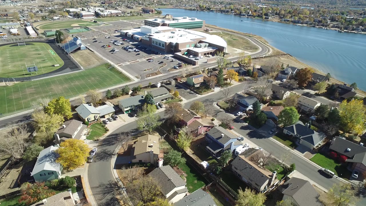 Second aerial frame of Hidden Lake, Colorado with rooftops and a curved street in the lower half, the athletic field on the left, and the lake behind a large shoreline building.