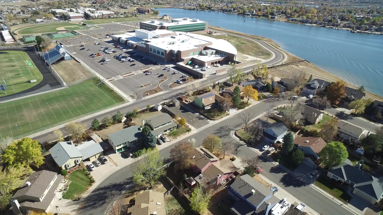 A slightly later aerial view of Hidden Lake, Colorado keeps the sports fields on the left, neighborhood roofs below, the large building and parking lot centered, and no visible people.