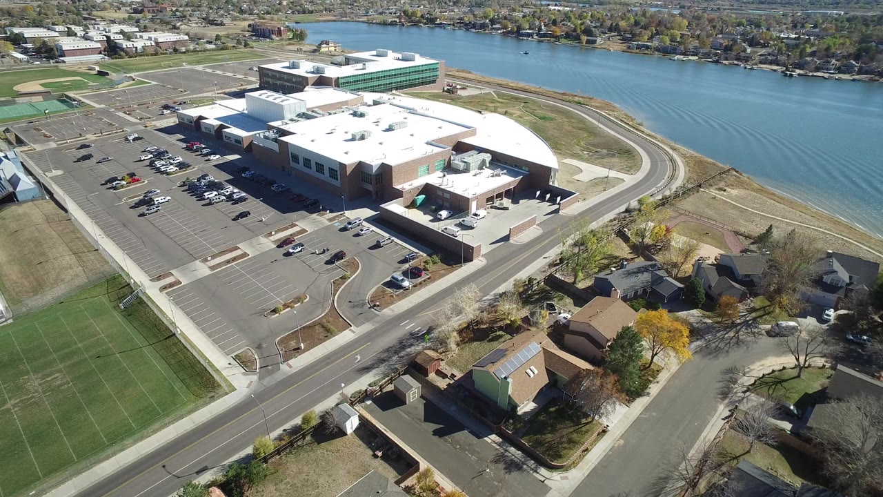 Aerial view of a large building complex beside a lake, parking lots, roads, fields, and homes.