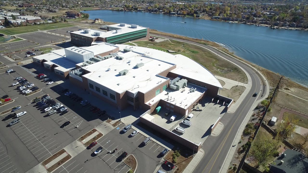Opening aerial frame over a white-roofed business or shopping complex in Hidden Lake, Colorado, with parked cars, a curved road, blue water behind it, and no visible people.