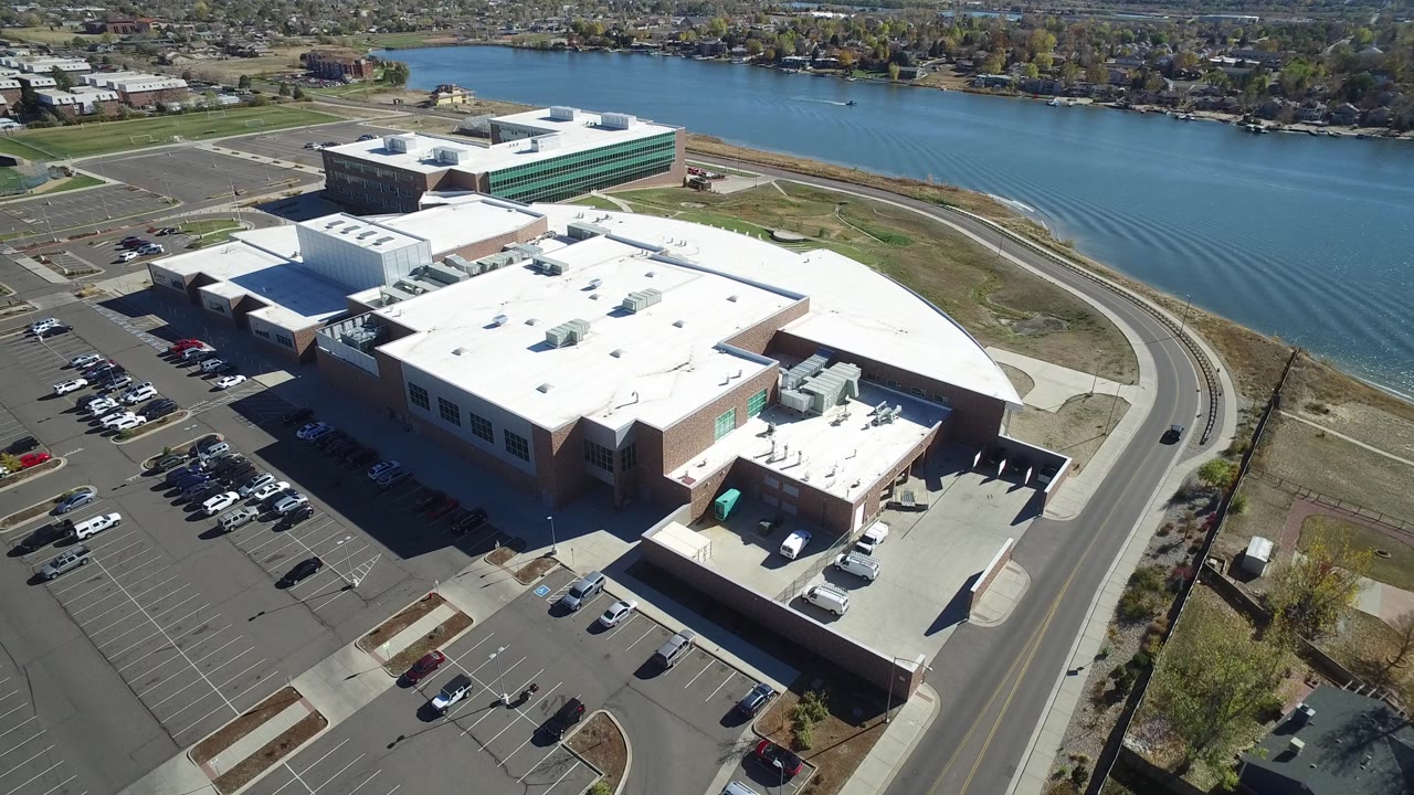 Aerial frame in Hidden Lake, Colorado, looking across the complex roof toward the water body, with parking rows at left, a curved road at right, and no visible people.