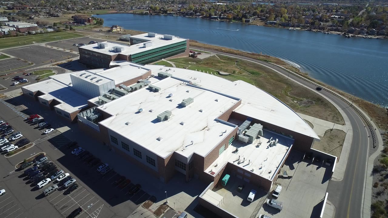 Aerial view of a large flat-roofed building beside a lake and parking lots.