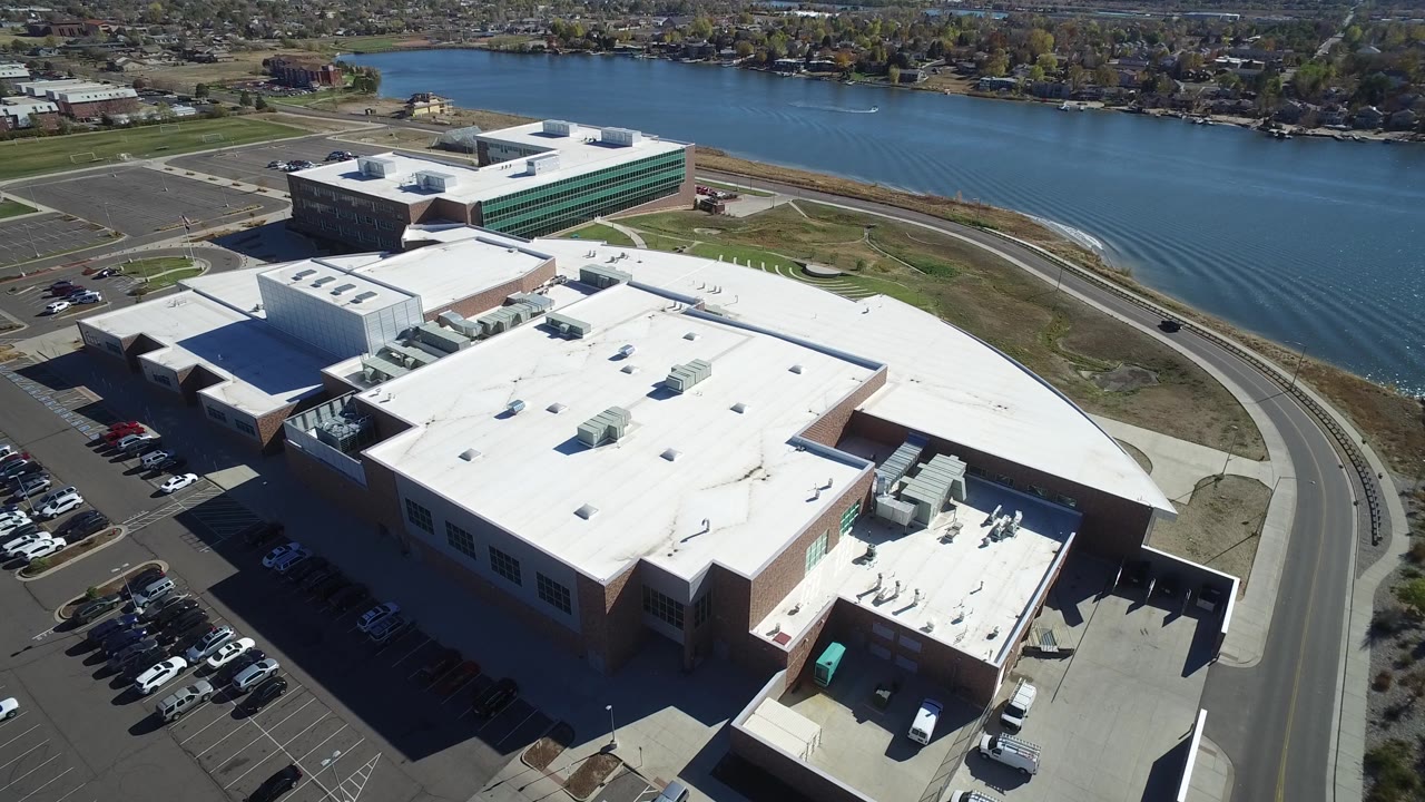 Aerial view in Hidden Lake, Colorado showing more of the central white roof, adjacent parking rows, a curved lakeside road, blue water, far residential blocks, and no visible people.