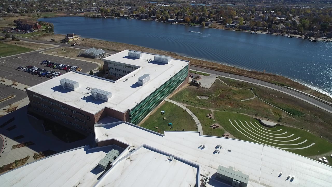 Aerial view of a white-roofed building beside blue water and a parking lot.