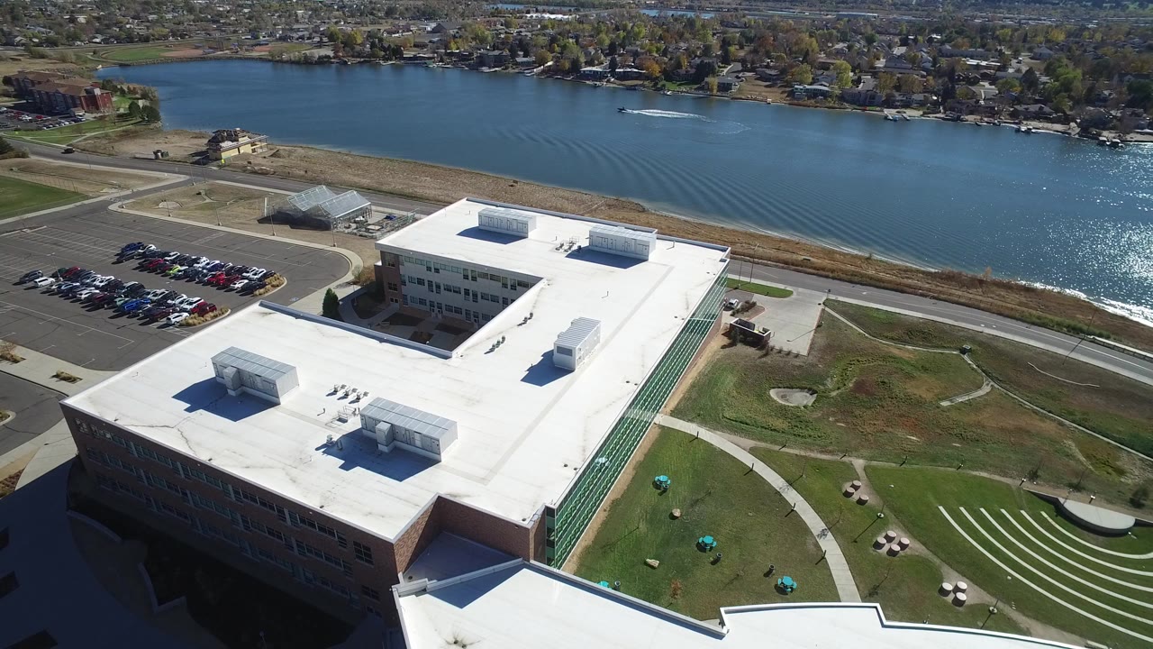 Aerial view of a large white-roofed building beside a road and lake.