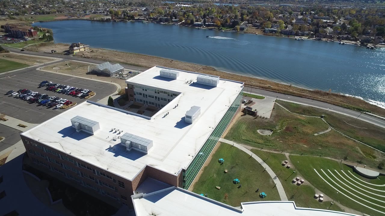 Aerial view in Hidden Lake, Colorado, with the building roof centered lower in frame, a parking lot to the left, road by the shoreline, lake water, and no visible people.