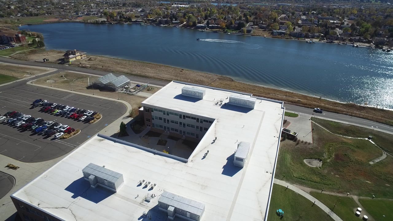 Aerial view in Hidden Lake, Colorado showing a large white-roofed building in the foreground, parked cars at left, a shoreline road, blue lake water, and homes beyond the lake.