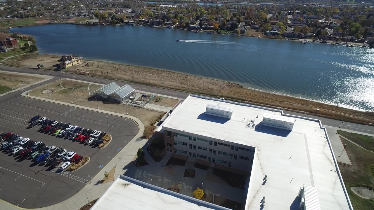 High aerial frame over Hidden Lake, Colorado, showing a white-roofed commercial building, parked cars, a road, and the blue lake beyond with no visible people.