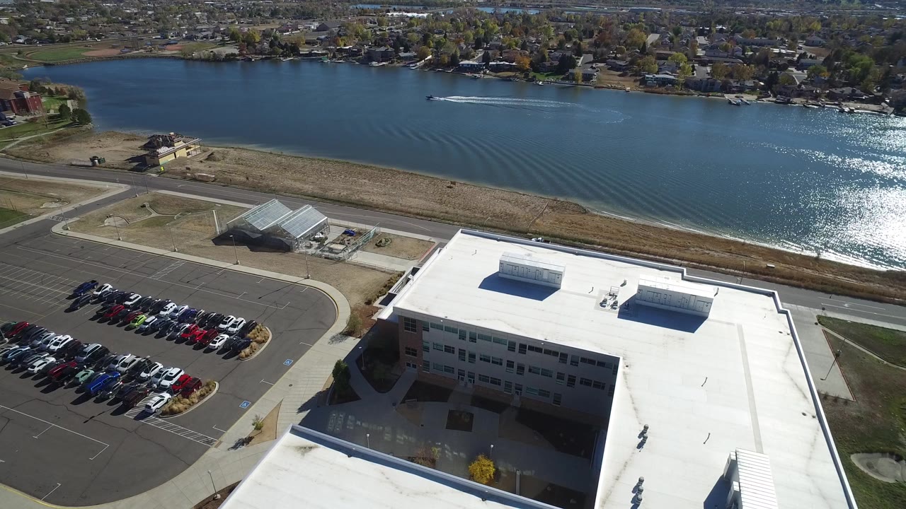 Aerial view in Hidden Lake, Colorado, with rows of parked cars at left, a flat white roof at lower right, and sun glare on the lake in the distance with no visible people.