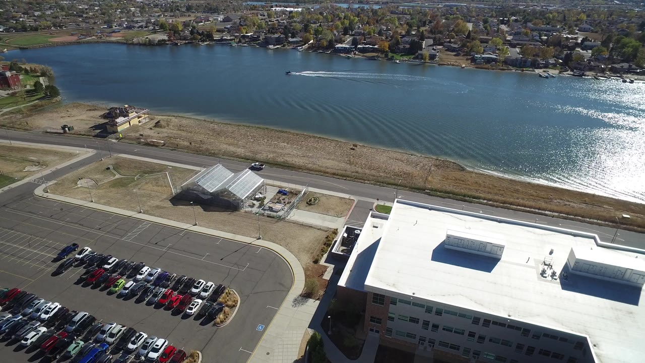 Aerial frame in Hidden Lake, Colorado, showing the lake with a wake line, dry shoreline, road, parking rows, a large white roof, and no visible people.