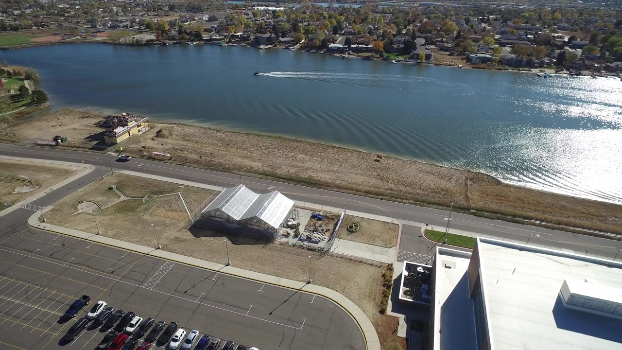 At 0:01, the speed boat has advanced across Hidden Lake, Colorado, leaving longer wake lines while the foreground road, parking spaces, dry grass, and white-roofed building remain visible with no visible people.