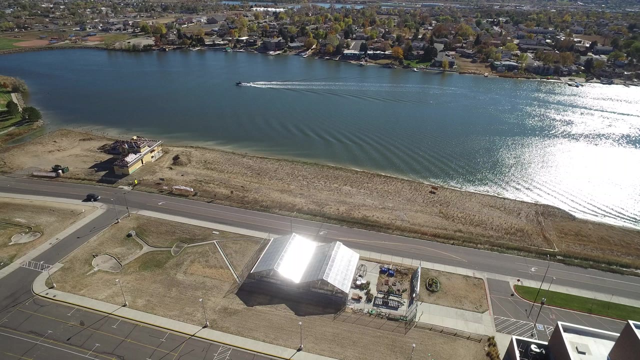 Aerial frame over Hidden Lake, Colorado showing blue-green water, a bright sun reflection near the right side, a shoreline road and open grass in the foreground, houses along the far bank, and no visible people.