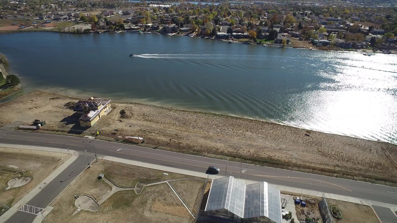 Aerial frame over Hidden Lake, Colorado, showing the boat farther across the lake, a curved wake trailing behind it, bright reflected light on the water, the roadside foreground, and no visible people.