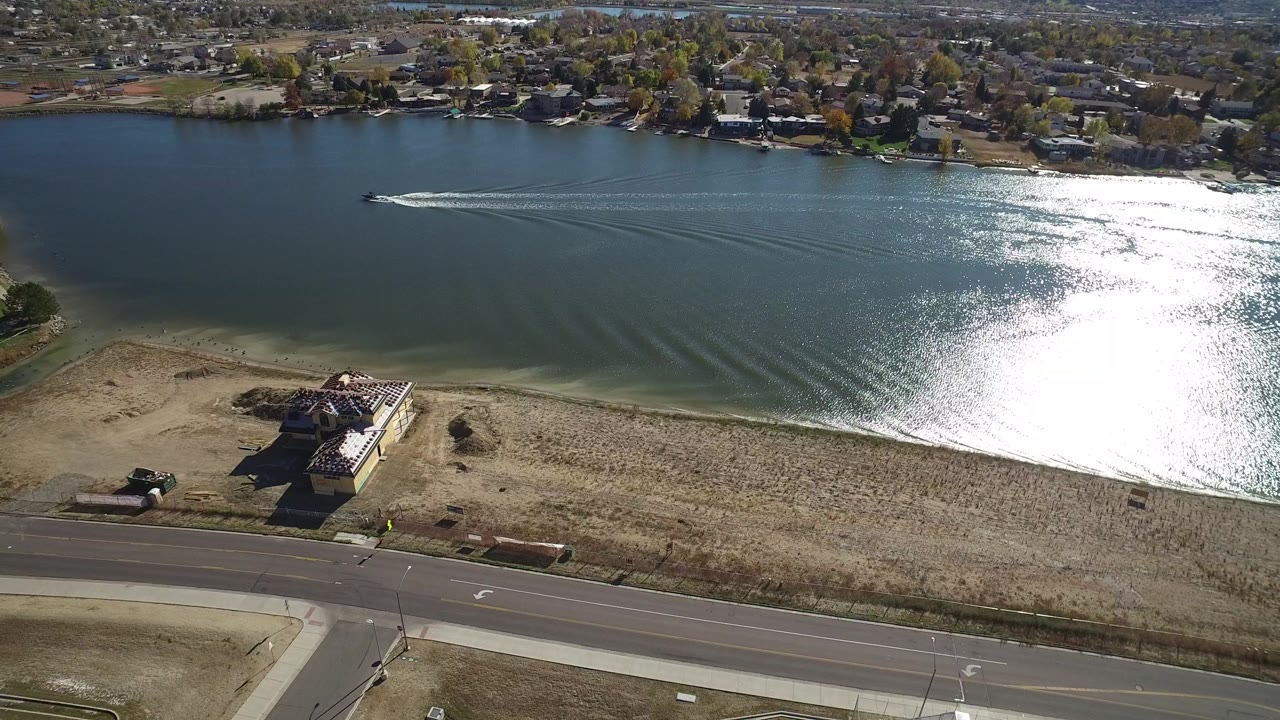 Aerial view of a lake with shoreline road, waterfront house, and boat wake.