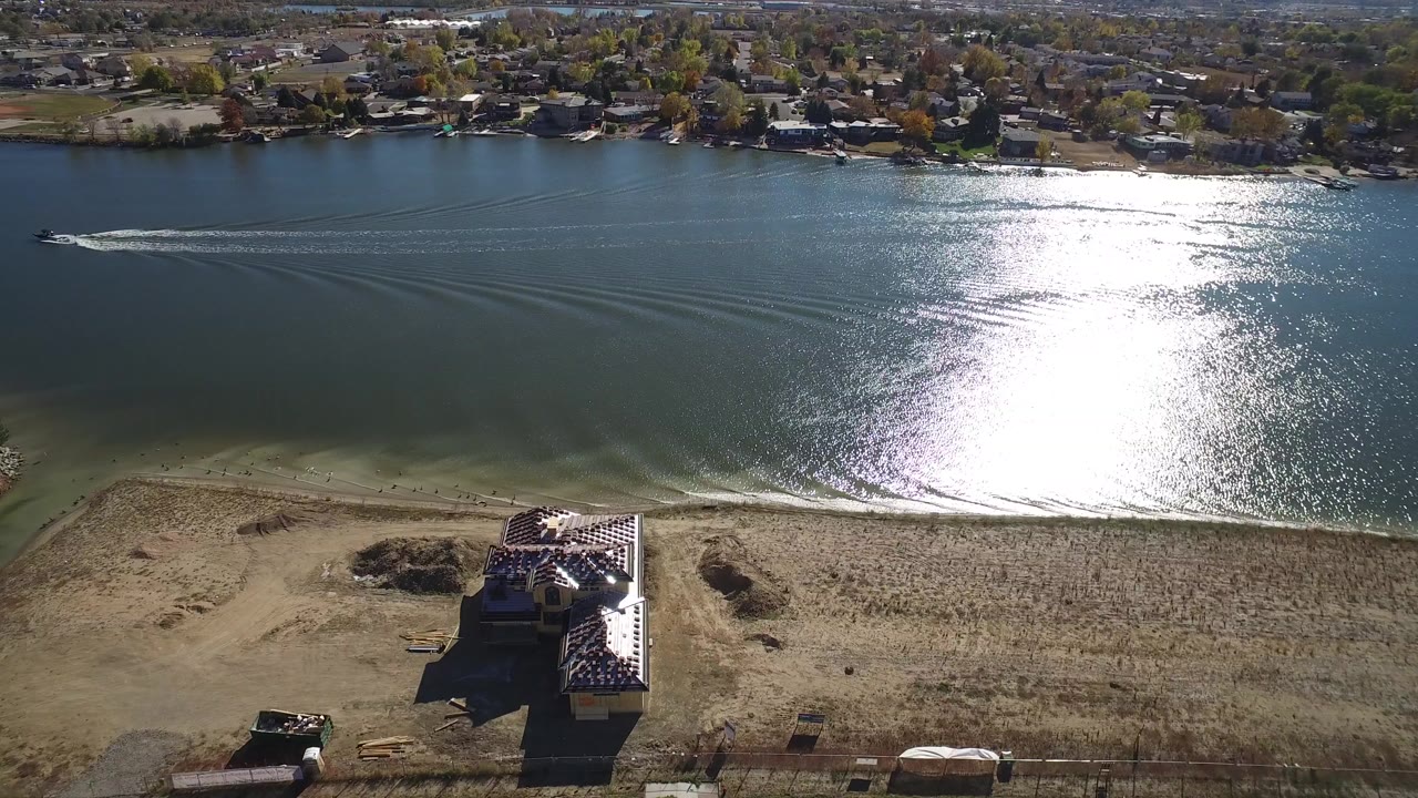 Aerial frame from Hidden Lake, Colorado showing a large home under construction between a paved road and bright water, with distant houses and no visible people.