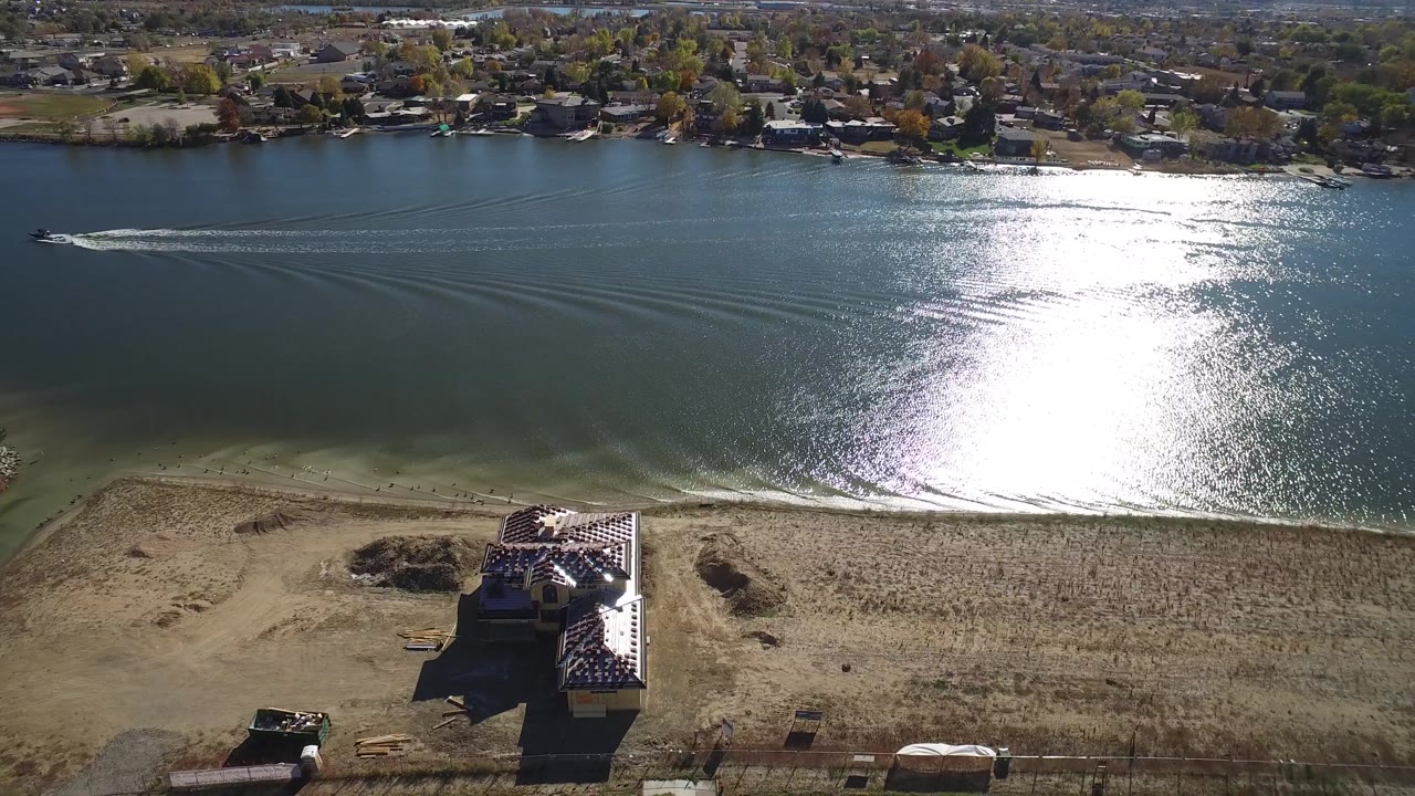 Aerial view in Hidden Lake, Colorado with the unfinished house lower in frame, a dumpster near the lot, wake lines crossing the water, and no visible people.
