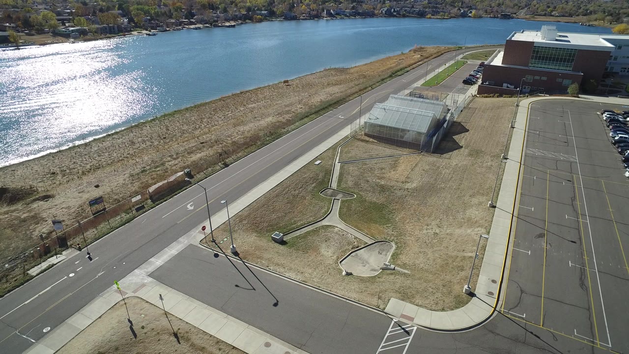 A high aerial frame over Hidden Lake, Colorado shows bright lake water, a shoreline road, dry grass, sidewalks, lampposts, parking lanes, a small shelter structure, a brick building, and no visible people.