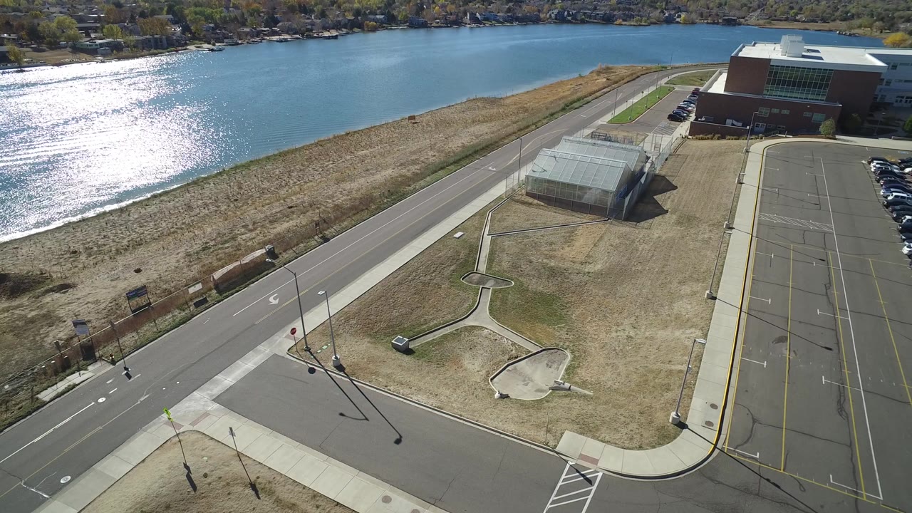 Drone-like overhead view of lake shoreline, road, sidewalks, dry grounds, and institutional building.