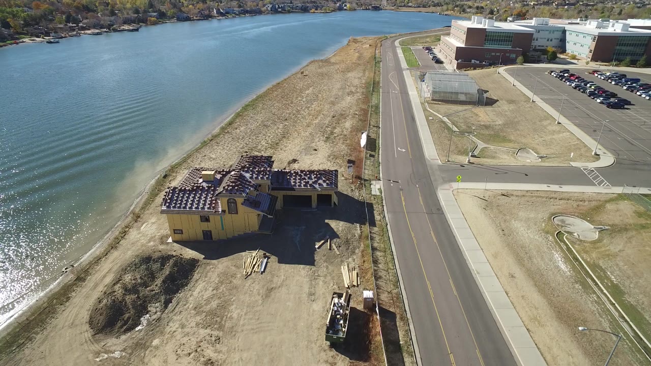 Aerial frame over Hidden Lake, Colorado showing blue water, a sandy shoreline, a yellow building under construction, a paved road, and larger buildings beyond, with no visible people.
