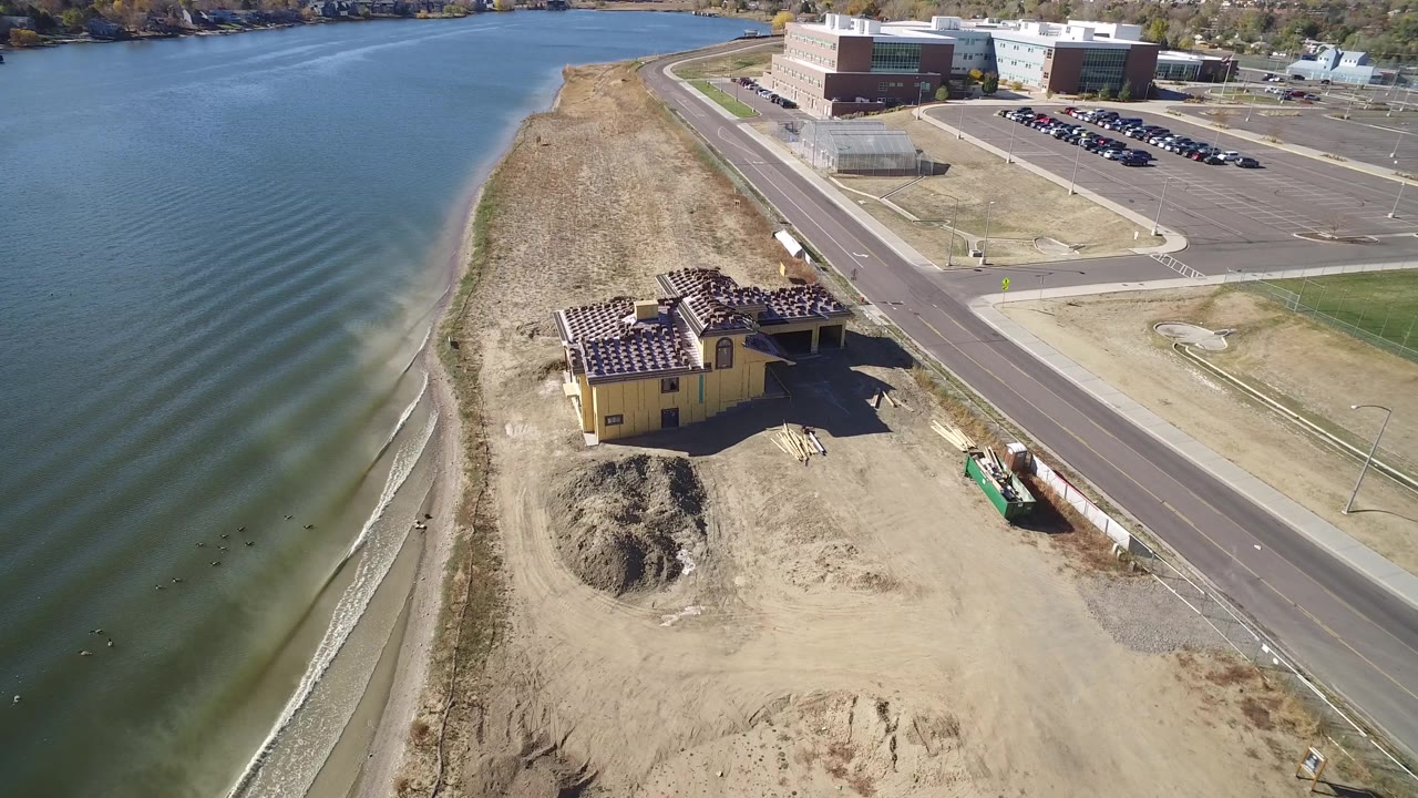 Aerial view of a lakeshore, road, yellow building, and construction dirt beside blue water.