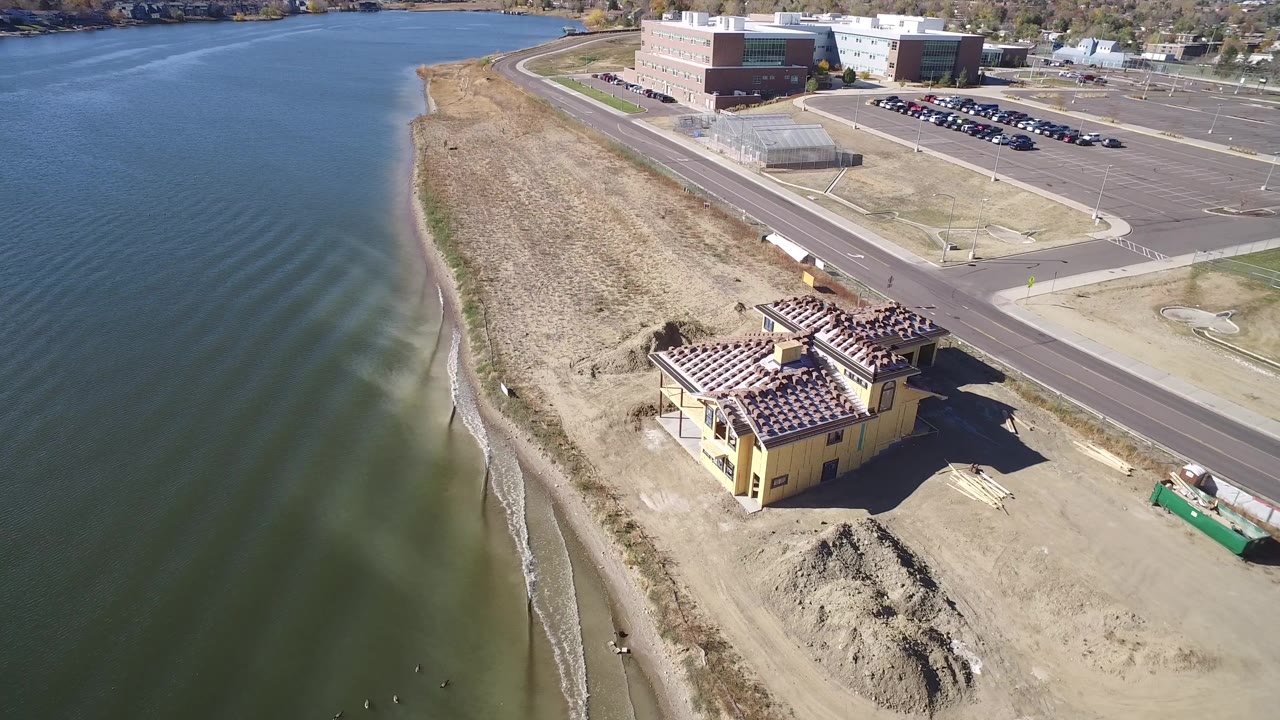 Aerial frame from Hidden Lake, Colorado showing green-blue water, a narrow sandy shoreline, a yellow construction building, dirt piles, a road, parking lots, and no visible people.