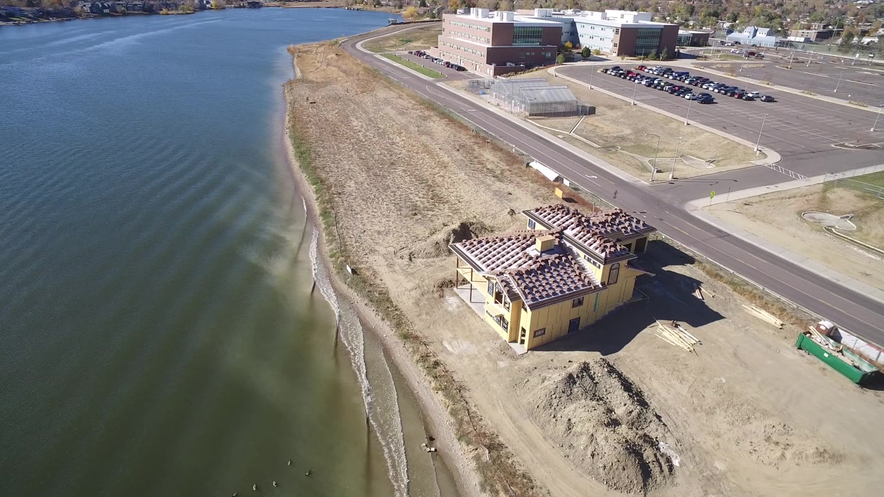 Aerial view over Hidden Lake, Colorado with rippled water filling the left side, a sandy beach strip, a yellow building under construction, road pavement, parking areas, and no visible people.