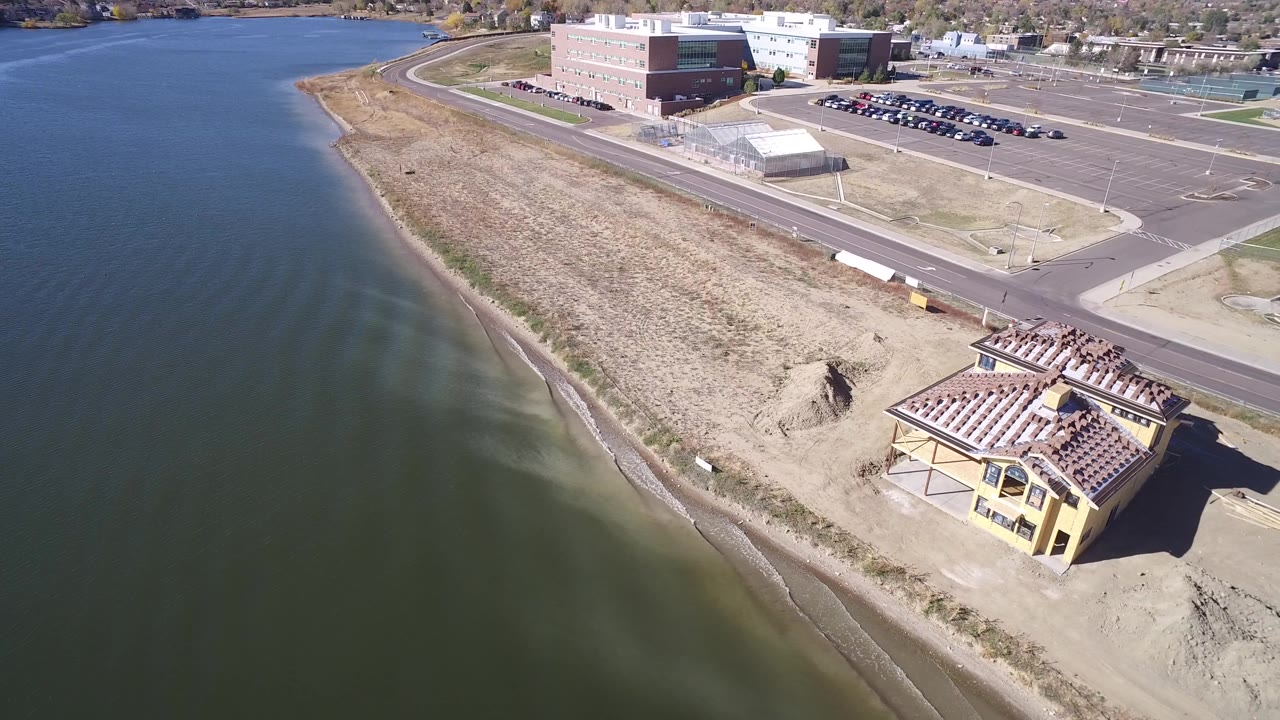 Aerial view of lake shoreline with a yellow building, road, parking lot, and larger buildings beyond.