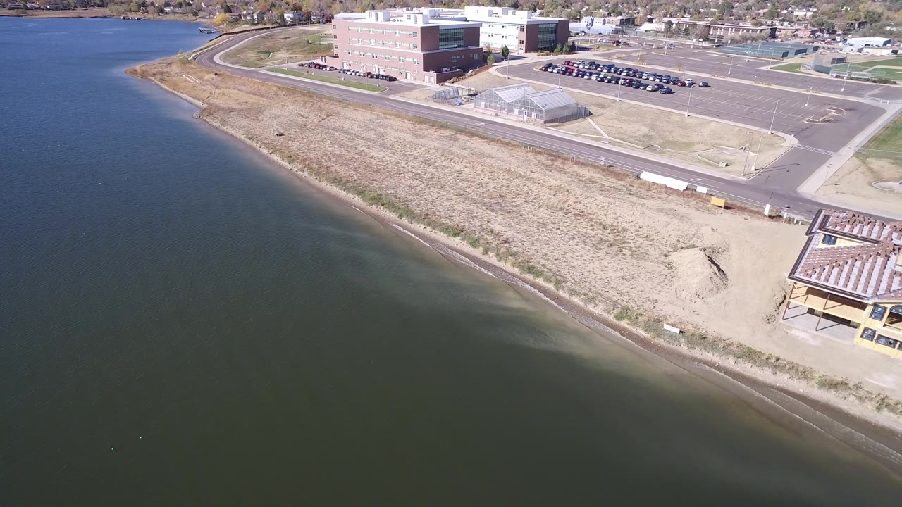 Aerial frame over Hidden Lake, Colorado, showing dark water, a narrow beach, dry grass, a road, parking areas, and large buildings in bright daylight, with no visible people.