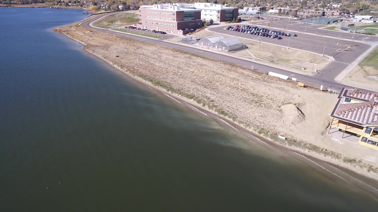 Aerial view of Hidden Lake, Colorado, with the waterline crossing the lower frame and a beach, grass strip, parking lot, and large blocky buildings beyond, with no visible people.