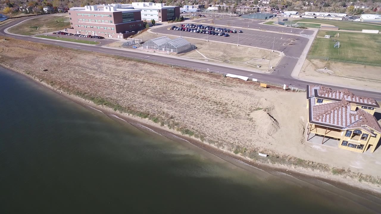 High aerial frame over Hidden Lake, Colorado showing dark calm water, a sandy beach, dry grass, a yellow beachside building, a road, and larger buildings beyond, with no visible people.