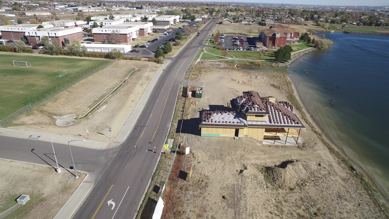 Elevated aerial frame over Hidden Lake, Colorado showing a paved road, dry grass, a roofed lakeside building, blue water, distant buildings, and no visible people.
