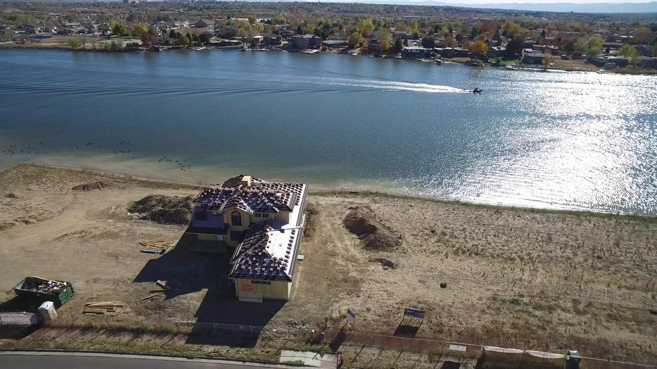 Hidden Lake aerial frame with the sun reflection centered on the water, a wake crossing the lake, a dry beach strip, a roadside building, and no visible people.