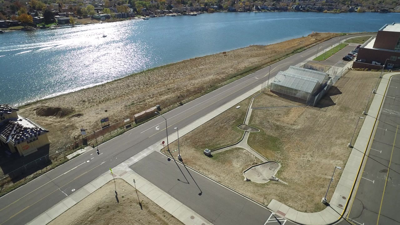 Opening aerial frame over Hidden Lake, Colorado, showing sunlit lake water, a shoreline road, a small parking area, dry grass, and distant homes with no visible people.