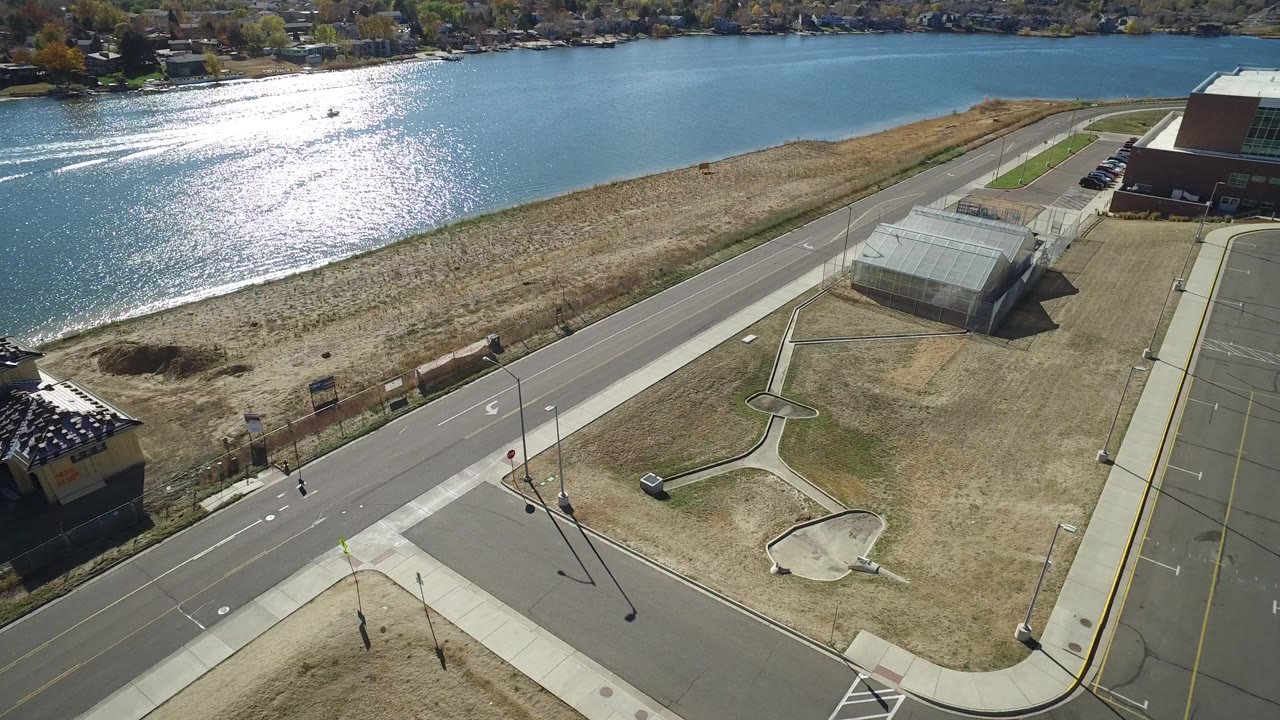 Drone view of a lakefront road, parking spaces, buildings, and dry shoreline grass.