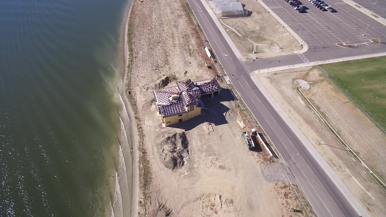 Overhead frame from Hidden Lake, Colorado showing green lake water, sandy shore, a single house under construction, road pavement, and parking areas, with tiny people visible near the shore.