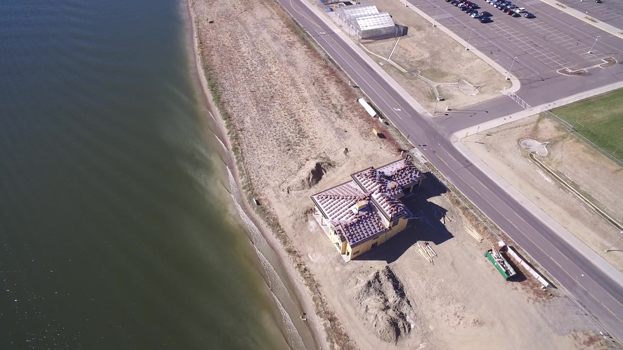 A slightly later overhead frame shows the empty Hidden Lake shoreline with the beach building lower in frame, parking lots at the top, road pavement to the right, and no visible people.