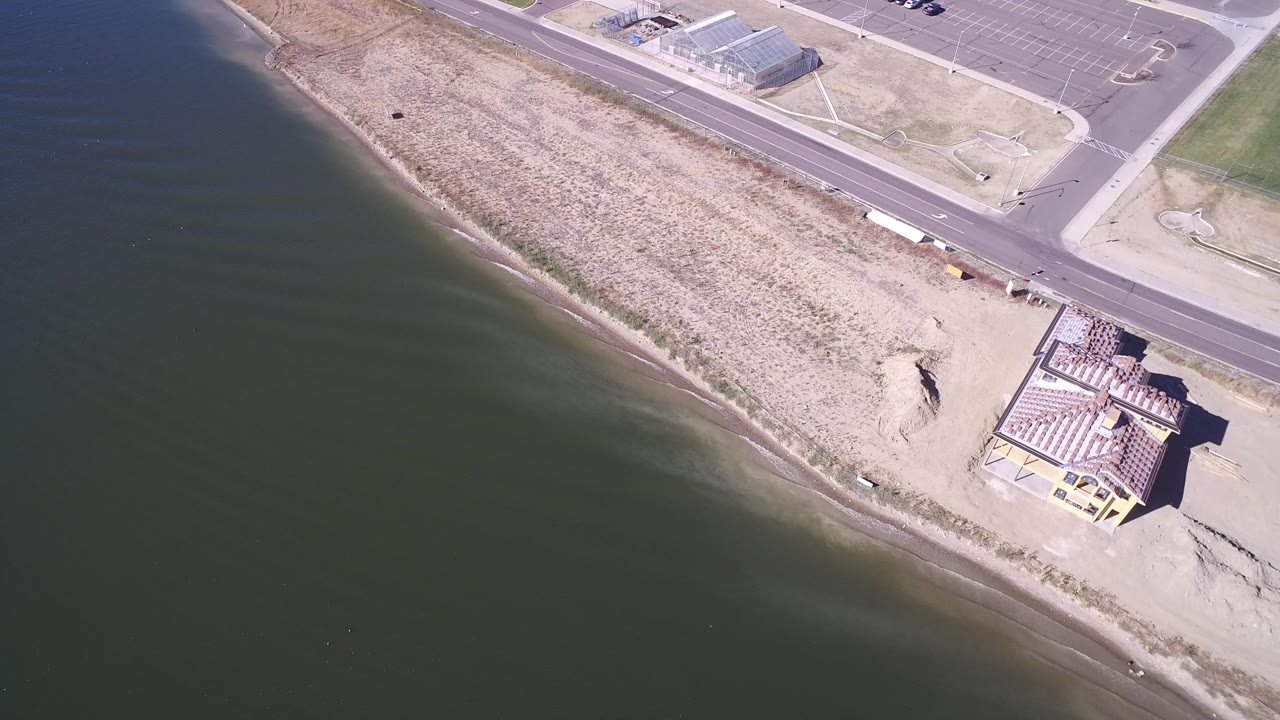 Aerial view of lake water, sandy shoreline, road, parking lot, and a roof-framed building.