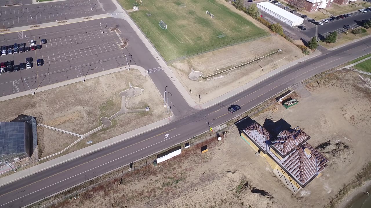 Drone-style overhead frame in Hidden Lake, Colorado with a dark car on the road, a mostly empty parking lot, green fields, a house by the water, and no visible people.
