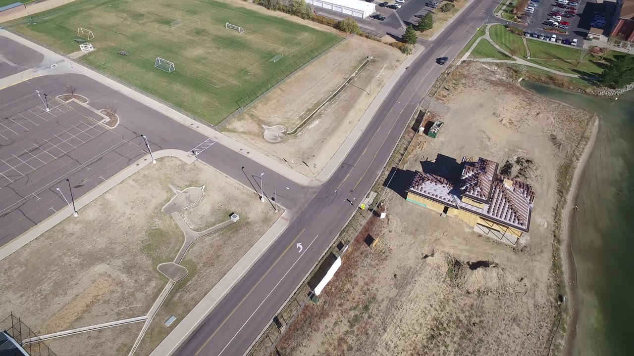 Opening aerial frame over Hidden Lake, Colorado showing a paved road between empty parking areas, dry grass, green soccer fields, a construction site by the water, and no visible people.
