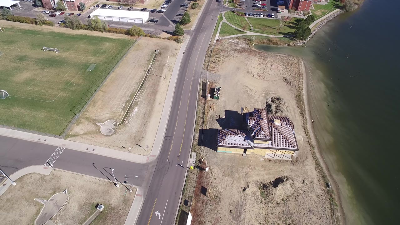 Aerial view in Hidden Lake, Colorado with a grass field and soccer goals at left, a road through the center, a lakeside construction site at right, and no visible people.
