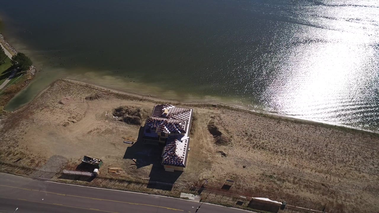 High aerial frame over Hidden Lake, Colorado, showing a shoreline house, dry grass, a paved road, sun glare on rippled water, and no visible people.