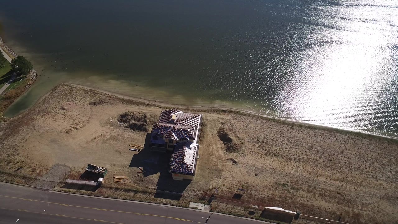 Aerial view in Hidden Lake, Colorado, with the house closer to center, bright reflection on the lake above it, road below, dry shoreline, and no visible people.