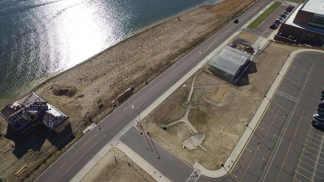 Aerial view of a road, empty parking lot, lakeshore, and nearby buildings.