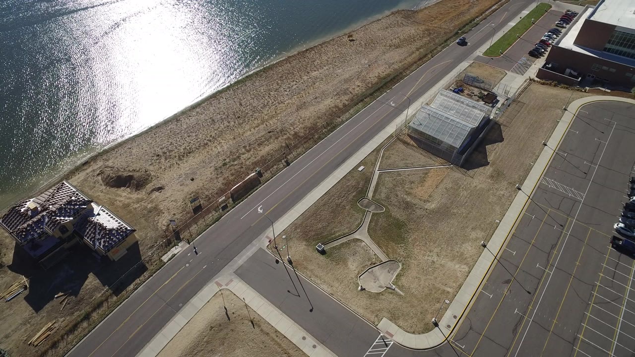 Aerial frame from Hidden Lake, Colorado with the road running between dry shoreline grass and a paved parking area, plus a building roof at lower left and no visible people.